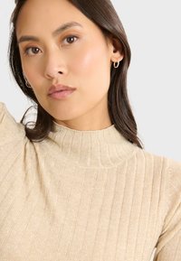 Beige Ripp-Turtleneck-Pullover mit strukturiertem Finish. Modell trägt kleine Creolen vor einem neutralen Hintergrund.