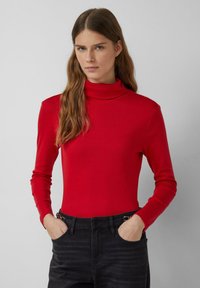 Junge Frau mit langen braunen Haaren, die einen roten Rollkragenpullover und schwarze Jeans trägt, steht mit den Händen in den Taschen vor einem grauen Hintergrund.