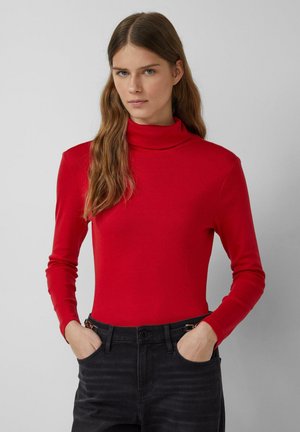 Junge Frau mit langen braunen Haaren, die einen roten Rollkragenpullover und schwarze Jeans trägt, steht mit den Händen in den Taschen vor einem grauen Hintergrund.