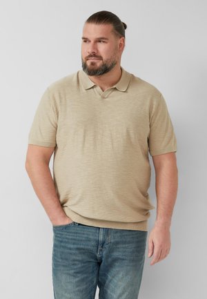 Mężczyzna z brodą i związanymi do tyłu włosami, ubrany w beżową koszulkę polo z krótkim rękawem i niebieskie dżinsy, stoi z jedną ręką w kieszeni, patrząc w bok.