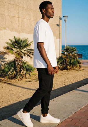 Homme en t-shirt blanc, pantalon noir et baskets blanches marchant le long d’un chemin au bord de la plage avec des palmiers et l’océan en arrière-plan.
