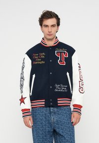 Chaqueta varsity azul marina y blanca con acentos rojos, que presenta texto y logotipos bordados. Cuello y puños acanalados, detalle de estrella en la manga.