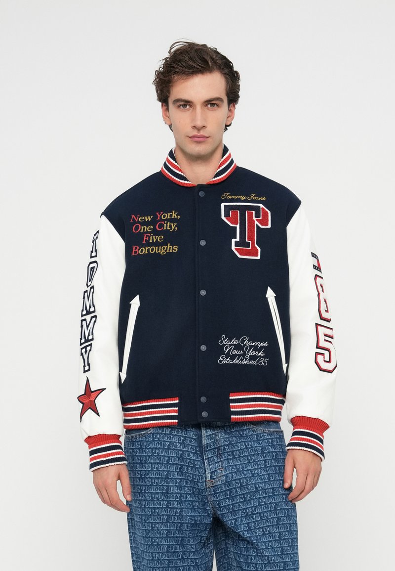 Chaqueta varsity azul marina y blanca con acentos rojos, que presenta texto y logotipos bordados. Cuello y puños acanalados, detalle de estrella en la manga.