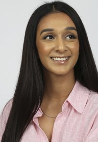 Jonge vrouw met lang donker haar, gekleed in een roze gestreepte blouse met kraag en een dunne zilveren ketting, die glimlacht tegen een neutrale achtergrond.