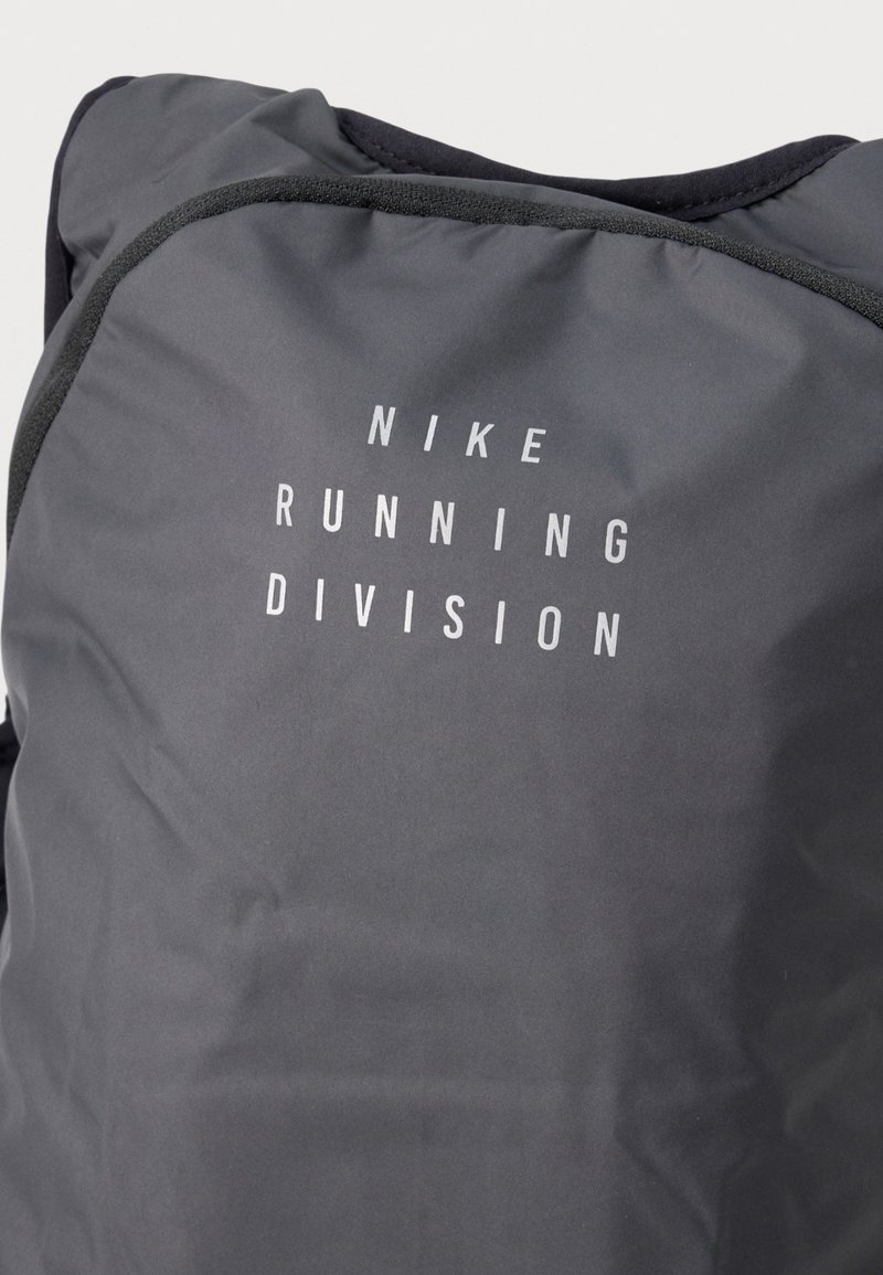Tissu gris foncé avec le texte blanc "NIKE RUNNING DIVISION" imprimé verticalement sur une surface rembourrée, probablement un sac ou une veste.