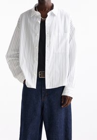 Personne portant une chemise blanche à manches longues à rayures verticales par-dessus un débardeur noir, un jean large foncé, une ceinture et un collier de perles.