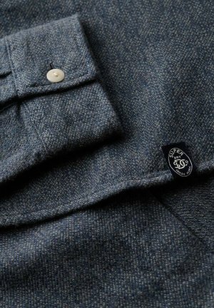 Tela texturizada gris oscuro con un puño abotonado blanco y una pequeña etiqueta negra que dice "Super Dry Co Trade Mark."