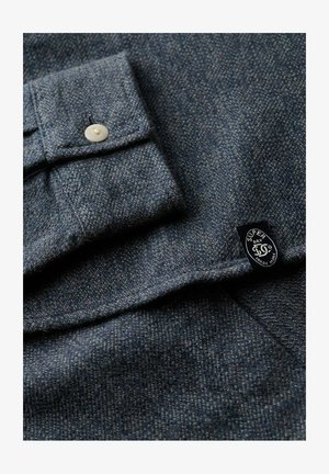 Tela texturizada gris oscuro con un puño abotonado blanco y una pequeña etiqueta negra que dice "Super Dry Co Trade Mark."