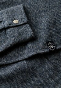 Close-up af tekstureret mørkegrå skjortekant med hvid knap og lille sort Superdry logo mærke på ærmet.