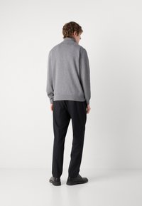 Muškarac stoji okrenut leđima, nosi sivu majicu s visokim ovratnikom, crne hlače i crne cipele, na jednobojnoj bijeloj pozadini.