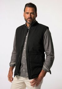 Chaleco sin mangas negro con cremallera frontal, que presenta dos bolsillos laterales y una textura suave, usado sobre una camisa de botones gris.