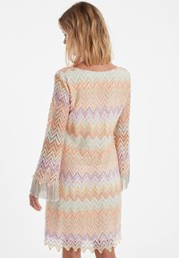 Robe en dentelle avec un motif en zigzag dans des couleurs pastel, dotée de manches évasées avec des détails en frange. Le matériau semble léger et texturé.