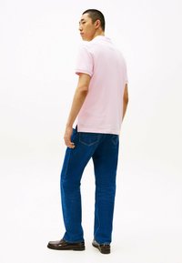 Lichtroze polo met korte mouwen, gecombineerd met blauwe denim jeans en zwarte instappers, vanuit de achterkant weergegeven tegen een witte achtergrond.