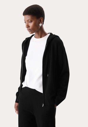 Vrouw met kort haar die zilveren oorbellen draagt, een wit overhemd, een zwarte zip-up hoodie en een zwarte broek, kijkt naar beneden links.