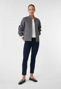 Grijze jas van een wolmix met een staande kraag, twee voorzakken en geribbelde manchetten over een witte gebreide top, gecombineerd met donkere spijkerbroeken en zwarte ballerina's.