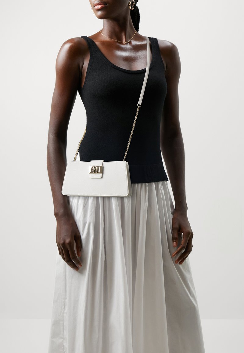 Weiße Lederhandtasche mit einem geometrischen Verschluss, akzentuiert mit einer goldenen Kettenriemen. Das Modell trägt ein schwarzes Tanktop und einen fließenden weißen Rock.