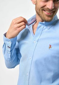 Világoskék gombos ing, gallérral és hímzett logóval. A gallér és az ujjak belsejében piros és kék csíkos díszítés található. 100% pamut anyag.