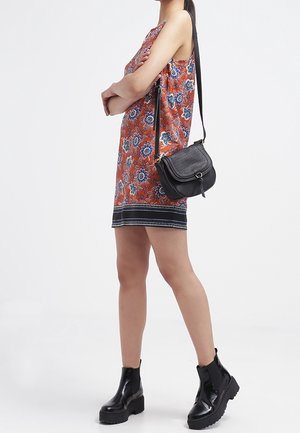 Donna che indossa un vestito floreale arancione senza maniche, stivali neri fino alla caviglia e una borsa a tracolla nera, in piedi con le braccia incrociate.