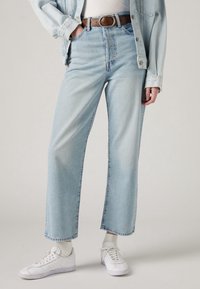 Vaqueros de pierna recta de cordura azul claro con cintura alta, que presentan un tono de mezclilla desgastado, una bragueta con botones y un cinturón visible con una hebilla marrón. Combinados con zapatillas blancas.