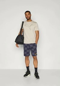 Ανδρική μπεζ μπλούζα Puma με γραφικό λογότυπο, σε συνδυασμό με ναυτικές κοντές παντελόνες με σχέδιο και μαύρα αθλητικά παπούτσια. Μια μαύρη τσάντα είναι κρεμασμένη στους ώμους.