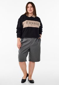 Svart och beige sweatshirt med texten "Firenze 1951" tryckt, kombinerad med grårandiga shorts. Modellen bär svarta platta skor. Svala tygtexturer.