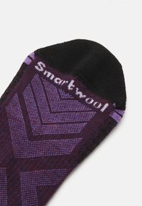 Lila och svart strumpa med chevronmönster, tillverkad av texturerad tyg. "Smartwool"-logotyp i vitt på den övre kanten. Närbild.