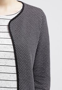 Grå texturerad cardigan med ett bikakesmönster, kantad i svart, bärs över en randig skjorta i en ljus färgpalett.