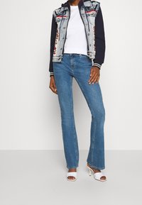 Veste en denim avec des détails brodés, t-shirt blanc, jean flare bleu clair et sandales à talons blancs ouvertes, présentés sur un fond uni.