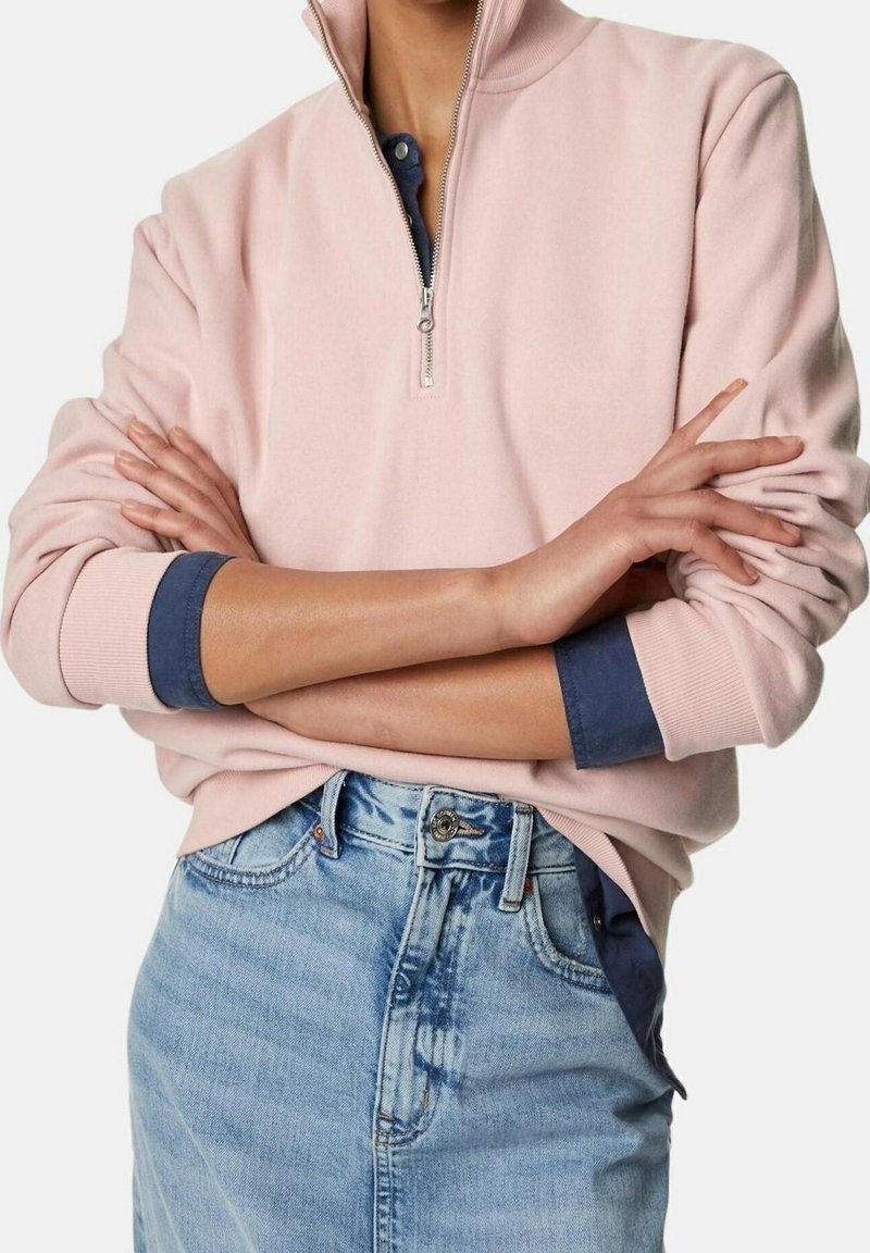 Lichtroze sweatvest met ritssluiting, hoge kraag en geribbelde boorden, gedragen over een donkerblauw shirt. Gecombineerd met een lichtblauwe spijkerrok.