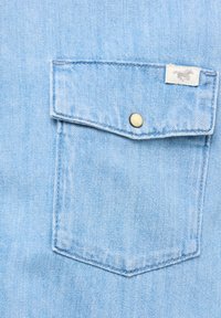 Tissu en denim bleu clair avec une poche poitrine dotée d'un rabat et d'un bouton-pression doré. La poche possède une petite étiquette avec un logo de cheval.