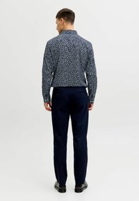 Mees seljaga vaataja poole, kannab tumel sinist mustrilist pika varrukaga särki, mereväe siniseid pükse ja musti kontsakingi, taustaks on lihtne valge taust.