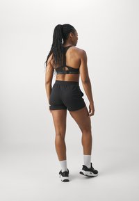 Sujeción deportiva negra con tirantes ajustables, combinada con pantalones cortos deportivos negros. La modelo lleva calcetas blancas y zapatillas de correr negras con acentos blancos.