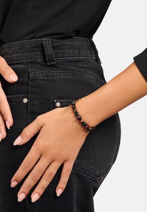 Perlenarmband mit abwechselnden Farbtönen aus schwarzen, grünen und braunen Steinen, getragen an einem Handgelenk mit hellem Hautton, das einen Kontrast zu dunkler Jeans bildet.