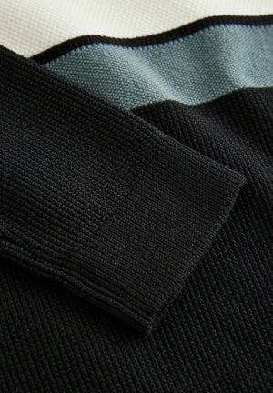 Tissu tricoté noir avec une texture côtelée, présentant une manchette noire se chevauchant des panneaux gris clair et blanc. Chaque partie a un contraste de couleur distinct.