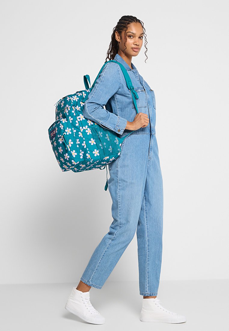 Mujer con mono de mezclilla y zapatillas blancas, llevando una mochila floral en tonos teal, de pie en perfil contra un fondo gris liso.