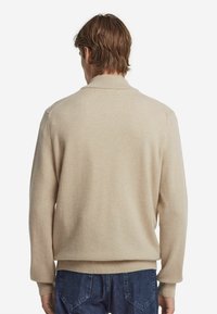 Beige stickad tröja med ribbad krage och ärmslut, med en avslappnad passform och mjuk textur. Bärs över blå denimjeans.