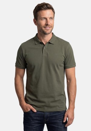 Mand med kort brunt hår iført en olivengrøn poloshirt og mørke jeans, stående med den ene hånd i lommen, kigger let mod højre.