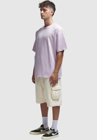 Lys lilla oversized t-skjorte, hvite cargo-shorts med sidelomme, og svarte sneakers med kremfargede detaljer. Enkelt design, avslappet stil.