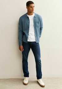 Ljusblå knapp-up kavaj över en vit t-shirt, kombinerad med mörka jeans och beige sneakers. Har en bröstficka och ribbade ärmslut.