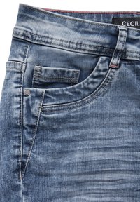 Jean en denim dans un bleu clair avec une texture légèrement déchirée. Comprend une poche avant, un bouton en métal et des coutures contrastées.
