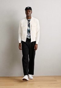 Chaqueta bomber blanca sobre una camisa de patrón multicolor, pantalones negros y zapatillas blancas, de pie contra un fondo sencillo.