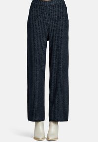 Högmidjade, vida mörkblå byxor med subtil vertikal randning, kombinerade med vita ankelstövlar på en enfärgad bakgrund.