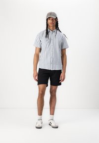Chemise à manches courtes, boutonnée, avec des rayures verticales bleues et blanches, associée à un short noir et une casquette grise, complétée par des baskets blanches.