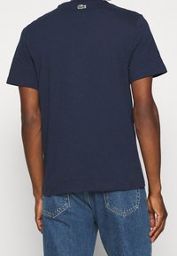 Muž v námořnickém modrém tričku s krátkým rukávem a modrých džínách, zobrazený zadečka s malým zeleným krokodýlím logem na límci trička.