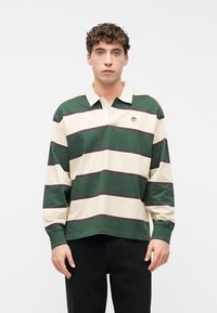 Νέος άνδρας που φοράει ριγωτό πουλόβερ ράγκμπι με φαρδιές πράσινες και μπεζ ρίγες και γιακά, μαζί με μαύρο παντελόνι, στέκεται μπροστά σε μονόχρωμο φόντο.