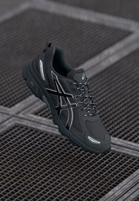 Black athletic shoe with a cushioned sole, mesh panels, and reflective accents. Features laces and a distinctive logo on the side.
