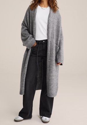 Femme portant un long cardigan gris tricoté par-dessus un t-shirt blanc, un jean noir à jambes larges et des baskets blanches, debout devant un fond uni.