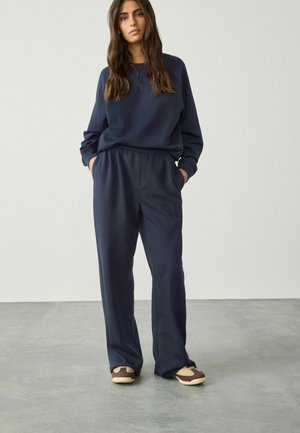 Navy blue sweatshirt with a round neckline and long sleeves, paired with loose-fitting navy sweatpants. The outfit is complemented by brown and cream shoes.