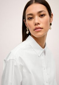 Hvid button-up skjorte med klassisk krave, glat tekstur og en tætsiddende design. Accentueret med store guld- og perleøreringe.