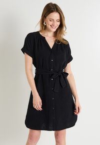 Vestido negro de botones con mangas cortas enrolladas, cintura ceñida y tela texturizada. Presenta un escote en forma de V y un ajuste holgado.